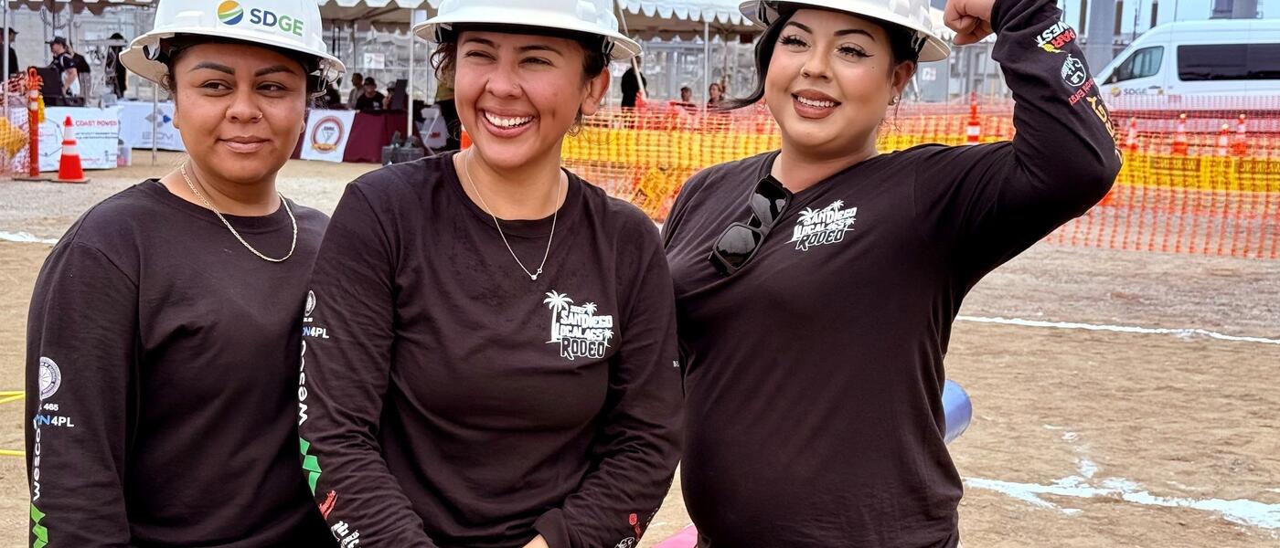 Three smiling women wearing white SDG&E hard hats pose for a photo.