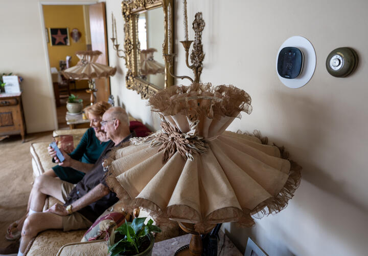 A modern thermostat alongside an older one, with a man and women talking in the foreground.
