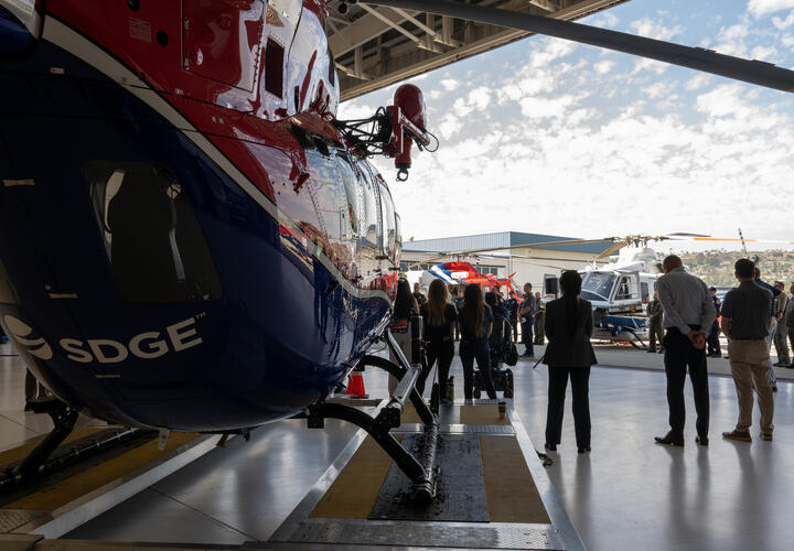 An image of a helicopter with the SDG&E logo on it, with a press conference taking place in the background.