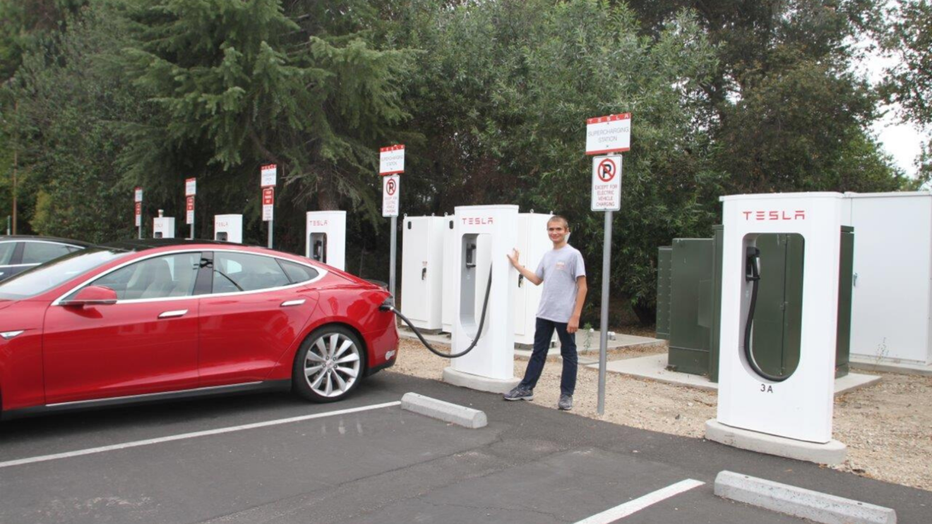 Brandon stands next to Randy's Tesla at a charging station.