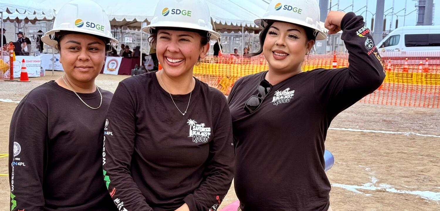 Three smiling women wearing white SDG&E hard hats pose for a photo.
