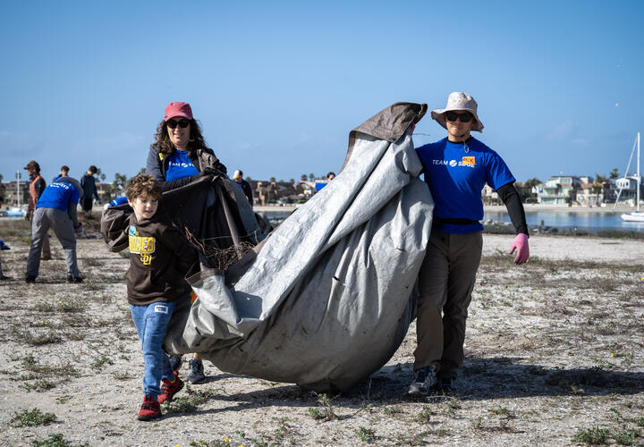 A young child, and two adults carry vegetation scraps in tarp. 