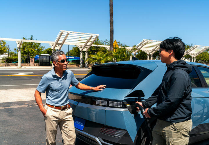 As two men talk, the younger of the two charges his electric vehicle. 