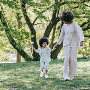 A woman holds a young childs hand, walking through a scenic setting with trees.
