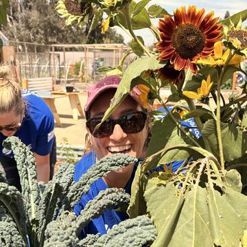 A smiling female volunteer pokes her head through garden greenery, with a vibrant sunflower positioned above her. 