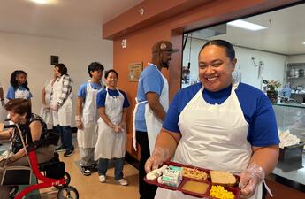 A woman wearing a white apron holds a tray of food.