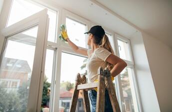 A woman with a ponytail stands on a ladder, cleaning the windows.
