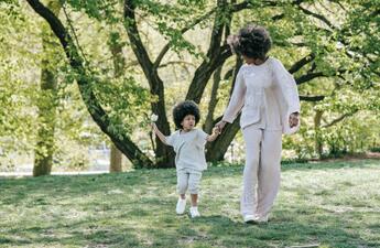 A woman holds a young childs hand, walking through a scenic setting with trees.
