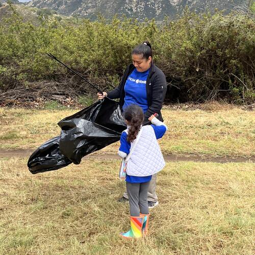 A woman wearing a bright blue shirt holds a trash bag as her daughter looks up at her. 